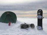 Snowy mountain camping setup with a tent, snowboard, and backpack in winter.