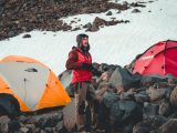 Man camping in snow-covered mountains with tents and rocky terrain.