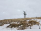 A coastal radar tower stands amidst snow-covered dunes and dry grass under a cloudy winter sky.