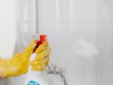 Crop unrecognizable person in yellow latex protective glove spraying detergent from bottle with plastic dispenser at glass of shower cabin
