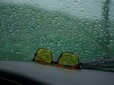 Yellow sunglasses rest on a car dashboard against a rain-speckled window.