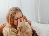 A woman sitting on a couch with eyes closed, holding her nose in apparent sinus pain.