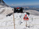 Snow groomer operating near avalanche warning sign in the Swiss Alps on a sunny day.