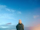 A person stands under a twilight sky, looking at the crescent moon. The scene captures a serene evening moment.