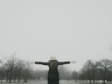 woman in black long-sleeved shirt standing and facing trees with stretched arms under white sky
