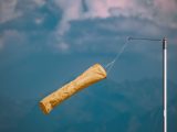 Yellow windsock blowing in the wind, mounted on a pole against a cloudy blue sky.