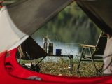 Through tent hole view of portable chairs and table with thermoses on grass shore against lake
