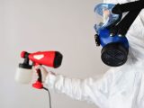 A worker in protective gear uses a disinfectant spray indoors.