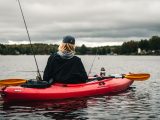Back view of a person in a red kayak fishing on a tranquil lake with a cloudy sky.
