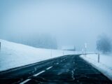 A foggy, snow-covered road stretches through a serene winter landscape, enveloped in mist.
