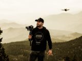 A man in a black hoodie photographing in misty mountains with a drone hovering.