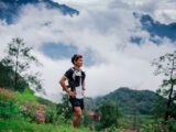 Trail runner pausing in lush green mountains with clouds and flowers.
