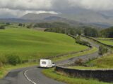 Car towing a caravan on a winding road through green hills in England.