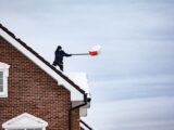 A man shoveling snow off the roof of a brick house during winter weather.