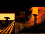 Silhouetted figures with umbrellas in a vibrant cityscape at night.