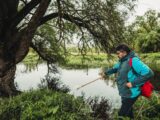 A man in outdoor gear exploring by a lake with lush trees during the day.