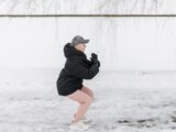 Senior woman doing squats outside in winter clothing on snowy ground.